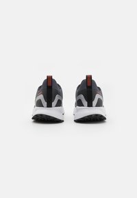 Des chaussures de sport avec une tige en mesh gris foncé, des accents noirs, une semelle intermédiaire blanche et une semelle extérieure texturée noire. Elles possèdent une languette de traction rayée de rouge.