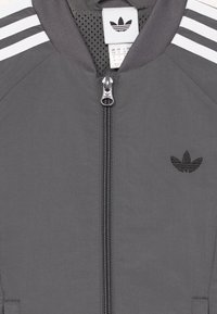 Sivá mikina Adidas so bielymi pruhmi na pleciach, predný zips s logom Adidas na ťahadle a logom Adidas trefoil na hrudi.