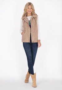 Beige Kunstfellweste mit hohem Kragen und Reißverschluss, kombiniert mit einem langärmeligen weißen Hemd, dunklen Jeans und hellbraunen Cowboy-Stiefeln.