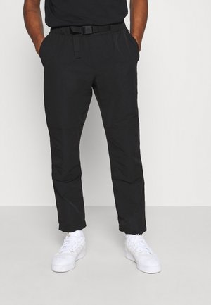 Persoon met zwarte taps toelopende broek met verstelbare riem en witte sneakers, staand met handen in de zakken tegen een effen achtergrond.