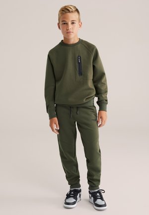 Felpa verde con tasca con zip, abbinata a pantaloni coordinati con cuciture laterali. Indossata con sneaker nere e bianche.