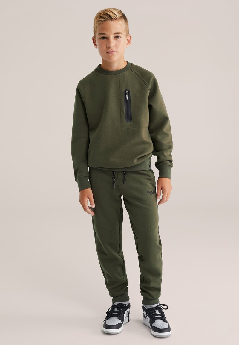 Zöld pulóver cipzáros zsebbel, összeillő nadrággal, amely oldalvarrással rendelkezik. Fekete-fehér sportcipővel viselve.