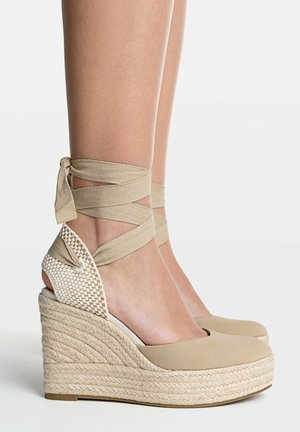 Beige Espadrille Keilabsatzschuhe mit Wickelriemen um die Knöchel an den Füßen vor einem schlichten weißen Hintergrund.