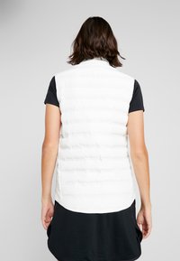 White sleeveless quilted vest with a high collar, featuring vertical seams and a longer back hem, worn over a black top and skirt.