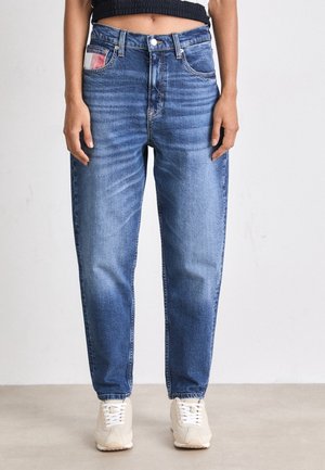 Kvinde iført højtaljede, afslappet blå jeans med falmede detaljer og lyse beige sneakers, stående mod en ensfarvet væg.