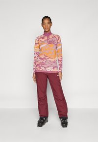 Woman standing wearing purple and orange patterned jacket, maroon pants, and black ski boots against plain background.