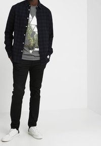 Marineblauwe geruite overhemd over een grijs T-shirt met geometrisch ontwerp, zwarte broek en witte sneakers. Strakke lijnen en casual fit.