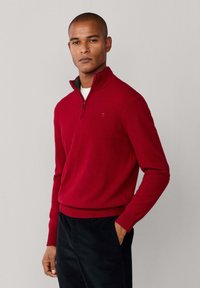 Roter gestrickter Pullover mit einem Viertel-Reißverschluss-Kragen, gerippten Bündchen und Saum. Getragen über einem weißen Hemd, das ein kleines Logo auf der Brust zeigt.