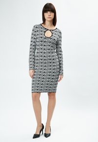 Schwarzes und weißes, gemustertes Bodycon-Kleid mit langen Ärmeln und einem Schlüssel-Ausschnitt-Design. Gestylt mit schwarzen High-Heels.