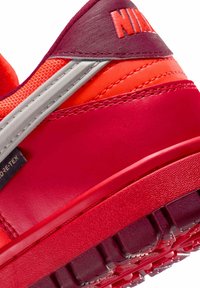 Rød og orange sneaker med en tekstureret gummisål. Har en glat finish, lagdelt design og reflekterende sølvaccenter.