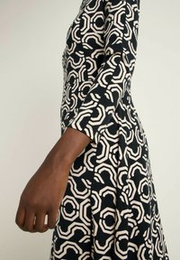 Schwarzes Kleid mit einem geometrischen cremefarbenen Muster, langen Ärmeln und einer taillierten Silhouette. Glatter Stoff mit leichtem Stretch.