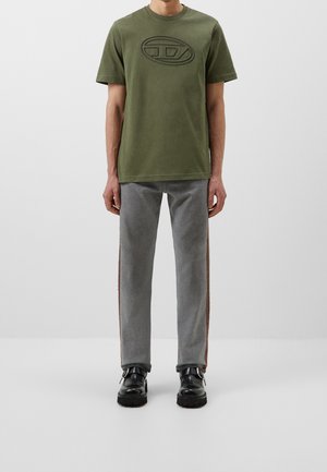 Man draagt een olijfgroen shirt met korte mouwen en een reliëflogo, grijze broek met bruine zijstrepen en zwarte leren schoenen, staand op een witte achtergrond.