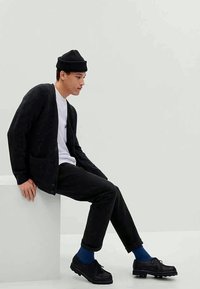 Černý pletený cardigan s předními kapsami, bílá košile, černé kalhoty, modré ponožky a černé boty s texturovanou podrážkou. Jednoduchý, moderní design.