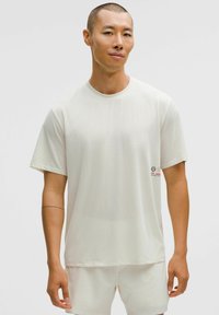 Chemise de sport légère à manches courtes, de couleur blanc cassé, avec un motif texturé et un petit logo sur le côté gauche. Tissu lisse.