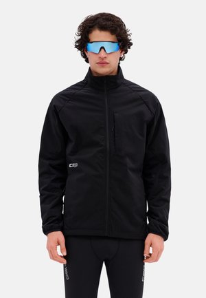 Mann trägt schwarze Jacke mit Reißverschluss und schwarze Leggings mit weißen Logos, trägt auch reflektierende blaue Sportsonnenbrille, steht frontal.