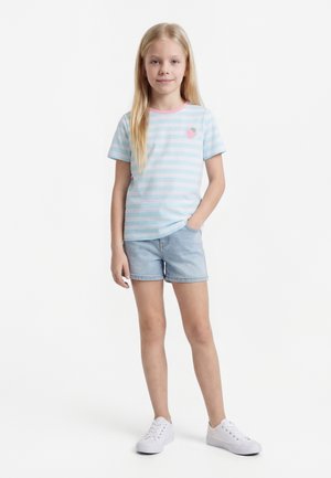 Young girl with long blonde hair wearing a light blue striped T-shirt with a pink strawberry patch, denim shorts, and white sneakers.