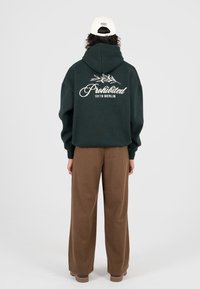 Persona che indossa un felpa verde scuro con la scritta "Prohibited 10119 Berlin" e un grafico di foglia, pantaloni larghi marroni e un cappellino bianco con la scritta "Modern Yuppies."