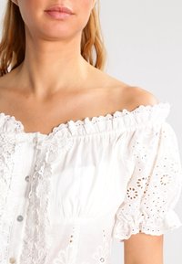 Žena oblečená v bílé blůze bez ramínek s krajkou a vyšíváním, krátkými nafouklými rukávy a knoflíky vpředu.