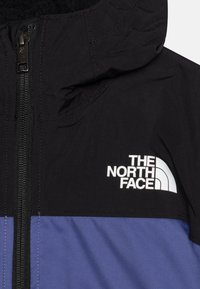 Musta ja violetti vetoketjullinen takki, jossa on huppu, teksturoitu kangas ja valkoinen "The North Face" -logo rinnassa.