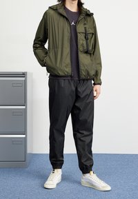 Olivgrön vattentät jacka med dragkedja och bröstficka, i kombination med svarta tajta byxor och vita sneakers, på en blå matta.