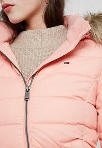 Jasnoróżowa puchowa kurtka z przednim zamkiem błyskawicznym, kapturem obszytym futrem, pikowaną strukturą oraz pod spodem beżowy sweter z dzianiny.