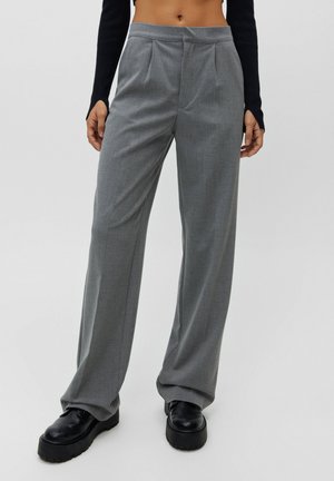 Das Model trägt eine locker sitzende graue Hose mit hohem Bund, kombiniert mit klobigen schwarzen Plateau-Schuhen und einem kurzen schwarzen Langarmtop, stehend vor weißem Hintergrund.