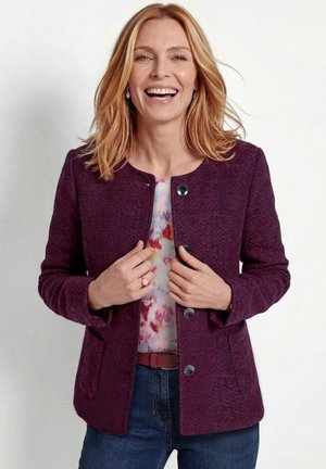 Femme souriante tenant le devant de sa veste texturée violette portée sur un chemisier coloré et un jean bleu sur un fond uni.