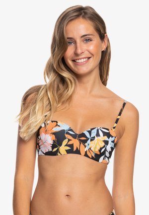 Young woman with long blonde hair smiles, wearing a black floral bikini top with orange, yellow, and blue leaves and flowers.