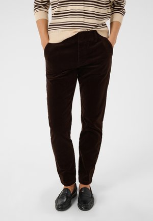 Personne portant un pantalon en velours côtelé marron foncé, des chaussures noires sans lacets et un pull beige avec des rayures horizontales marron, les mains dans les poches.
