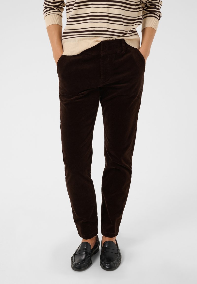 Personne portant un pantalon en velours côtelé marron foncé, des chaussures noires sans lacets et un pull beige avec des rayures horizontales marron, les mains dans les poches.