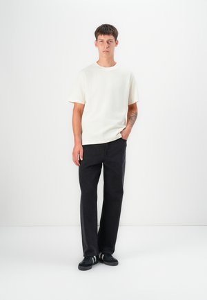 Hvid, ribbet t-shirt med korte ærmer, parret med sorte jeans med lige ben og sorte sneakers med hvide striber. Modellen står foran en ensfarvet baggrund.