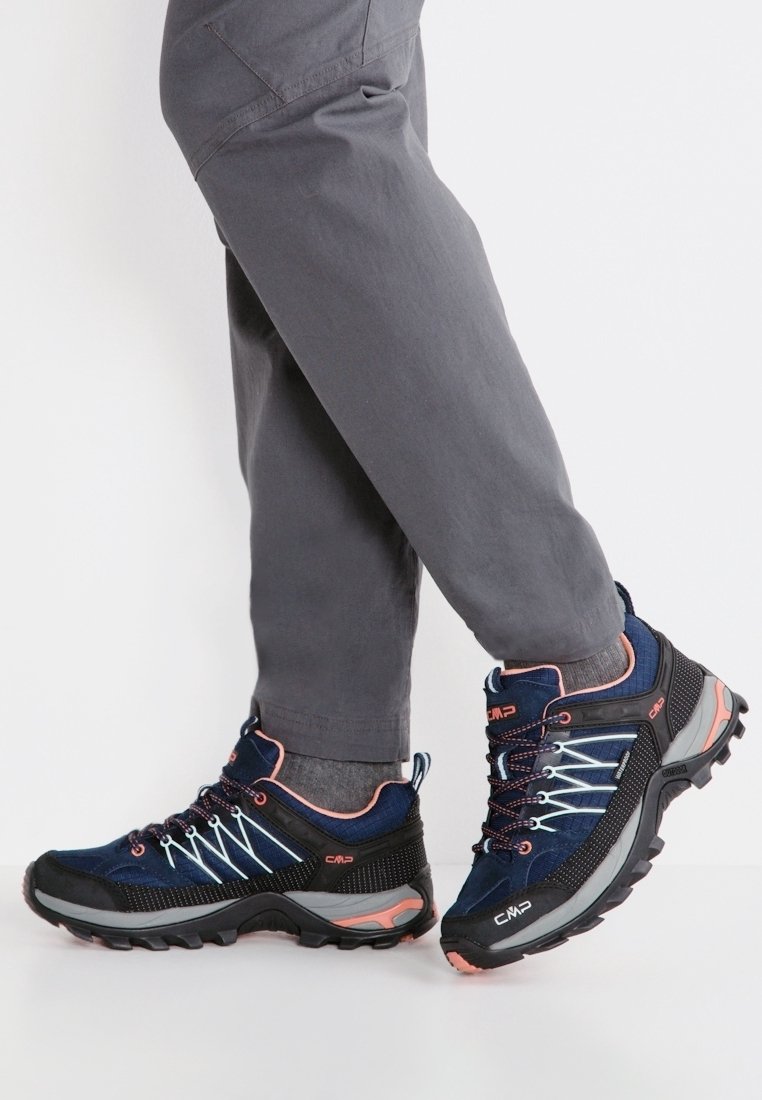 Osoba ubrana w szare spodnie i granatowe buty turystyczne z wytrzymałą podeszwą i różowymi akcentami, stojąca na białej powierzchni.