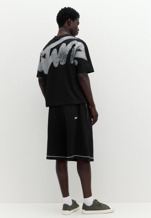 Homme debout face à un mur portant un t-shirt noir oversize avec un graffiti blanc et un short noir avec des baskets grises et des chaussettes blanches.