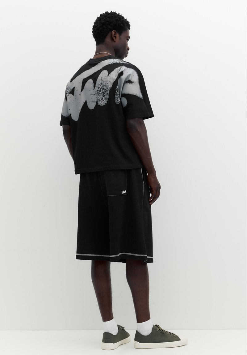 Homme debout face à un mur portant un t-shirt noir oversize avec un graffiti blanc et un short noir avec des baskets grises et des chaussettes blanches.