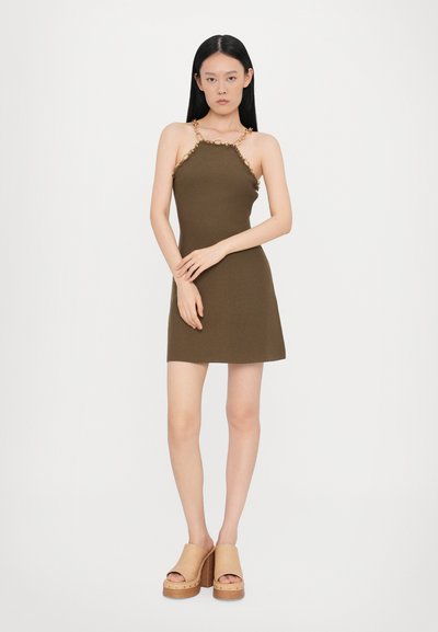 Olivgrünes ärmelloses Kleid mit tailliertem Oberteil und Kettendetails am Ausschnitt. Kombiniert mit beigen Plateausandalen mit hölzernem Akzent.