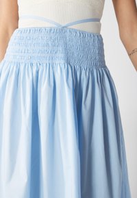 Jupe midi fluide bleu clair avec une taille smockée et un tissu doux. Présente des bretelles fines et ajustables à l'arrière.