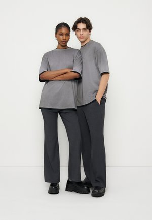 Deux personnes debout côte à côte portant des t-shirts gris surdimensionnés, des pantalons gris foncé et des chaussures à plateforme noires sur un fond blanc uni.