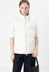 Hvit puffervest med høy krage, med glidelås foran og "Guess Jeans"-logo. Brukt over en lys topp, kombinert med mørke jeans og en svart veske.