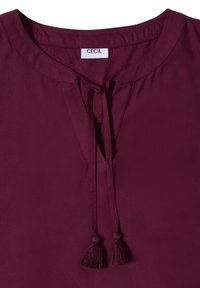 Blusa granate con escote redondo y lazo delantero con borlas, hecha de tela suave, con etiqueta de la marca "CECIL" dentro del cuello.