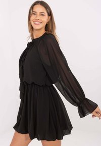 Czarna sukienka z lekkiego materiału, z długimi przezroczystymi rękawami, ozdobnym falbanowym przodem i elastyczną talią tworzącą rozkloszowany fason.