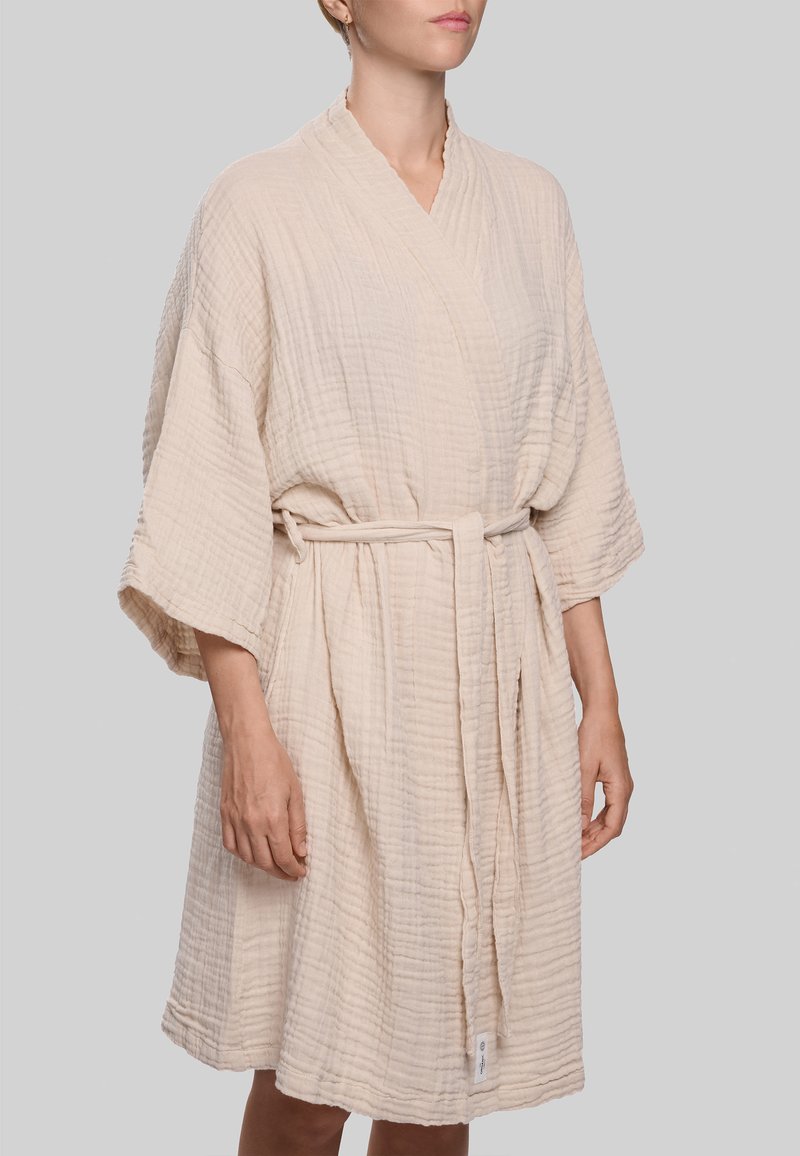 Laisvai krentanti smėlio spalvos chalatas iš tekstūruotos medžiagos, su apvyniojimo dizainu, plačiomis rankovėmis ir derančia juosta, surišta juosmens srityje.