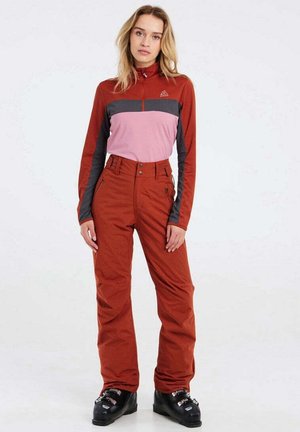 Rotes und pinkes Langarmoberteil mit einem grauen Streifen, kombiniert mit roten Hosen. Die Hosen haben eine hohe Taille und sichtbare Knöpfe. Schwarze Skistiefel.