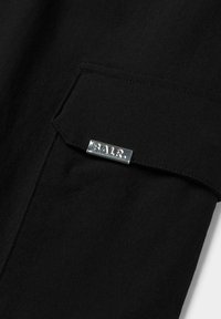 Sort stof med en glat tekstur har en lomme med et sølvmetalmærke, der er indgraveret med "BALR." Designet er minimalistisk og moderne.