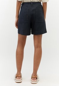 Pantalones cortos de lino azul marino con un corte relajado, que cuentan con dos bolsillos traseros y un dobladillo enrollado, combinados con sandalias de color claro.