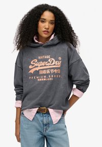 Sudadera gris carbón con gráfico frontal en durazno, que presenta un bolsillo canguro y un corte holgado. Usada sobre una camiseta rosa clara. Jeans de denim.