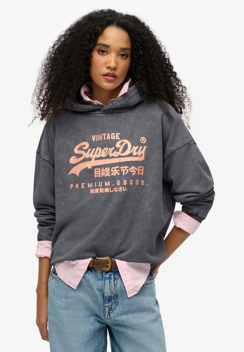 Sudadera gris carbón con gráfico frontal en durazno, que presenta un bolsillo canguro y un corte holgado. Usada sobre una camiseta rosa clara. Jeans de denim.