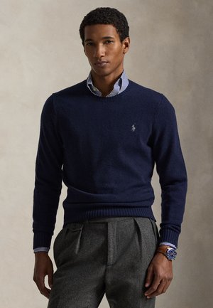 Άνδρας που φοράει μπλε πουλόβερ πάνω από ριγέ πουκάμισο, γκρι πλισέ παντελόνι και ρολόι χειρός με μπλε καντράν και καφέ δερμάτινο λουράκι.