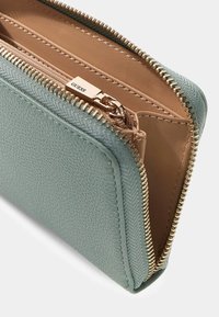 Grüne strukturierte Geldbörse mit beigem Innenfutter und goldfarbenen Beschlägen. Reißverschluss mit sichtbarem Markenschild im Inneren. Kompakte rechteckige Form.