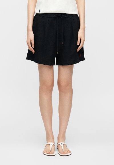 Γυναίκα που φοράει μαύρα σορτς με κορδόνι, λευκό τοπ με μικρό λογότυπο και λευκά παντόφλες, στέκεται μπροστά σε απλό φόντο.