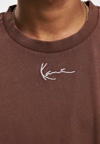 Brown cotton T-shirt with a ribbed crew neckline, featuring white embroidered signature on the chest area. Smooth texture.